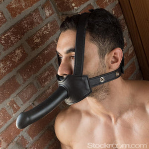 A shirtless man stands beside a brick wall wearing the black leather Head-On Gag Harness. The leather straps have metal snap closures.
