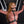 Load image into Gallery viewer, A woman with red hair in a mauve latex catsuit is restrained to a black St. Andrew’s Cross. She has the Silicone Bit Gag With Silicone Strap in her mouth and is wearing a black PVC bust harness. Various black wrist cuffs hang on the wall behind her.
