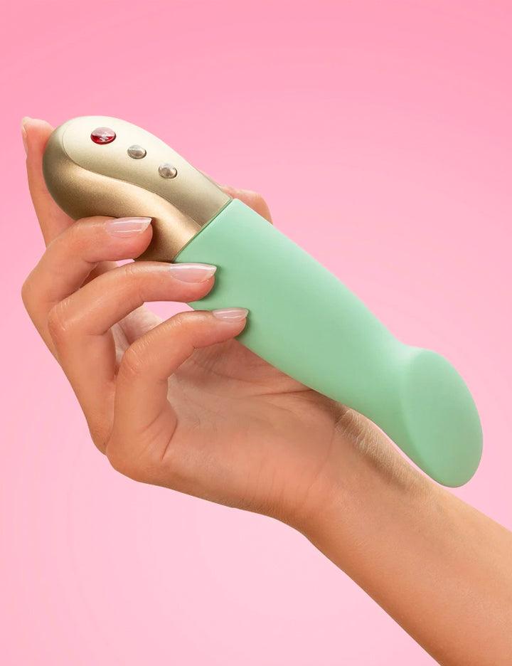 Somebody’s hand is shown holding up the Fun Factory Sundaze Pulsating Vibrator in Pistachio against a pink background.