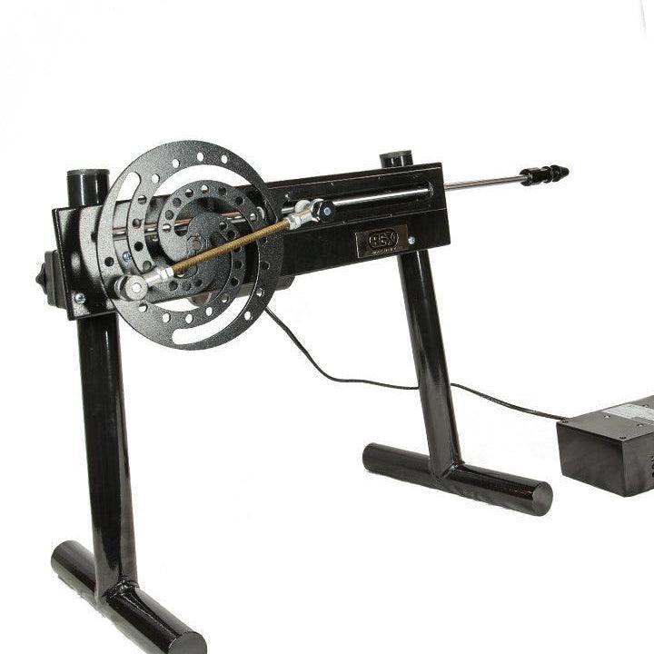 The Rex Machine is displayed against a blank background. It is a sex machine made of black metal. It is elevated on two legs and has a spiral wheel at the back with a gear attached leading to a pole. There is a grey control box next to it.