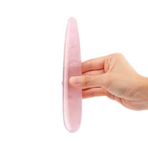 A hand is shown holding up a Le Wand Crystal Slim Wand made of Rose Quartz against a blank background.