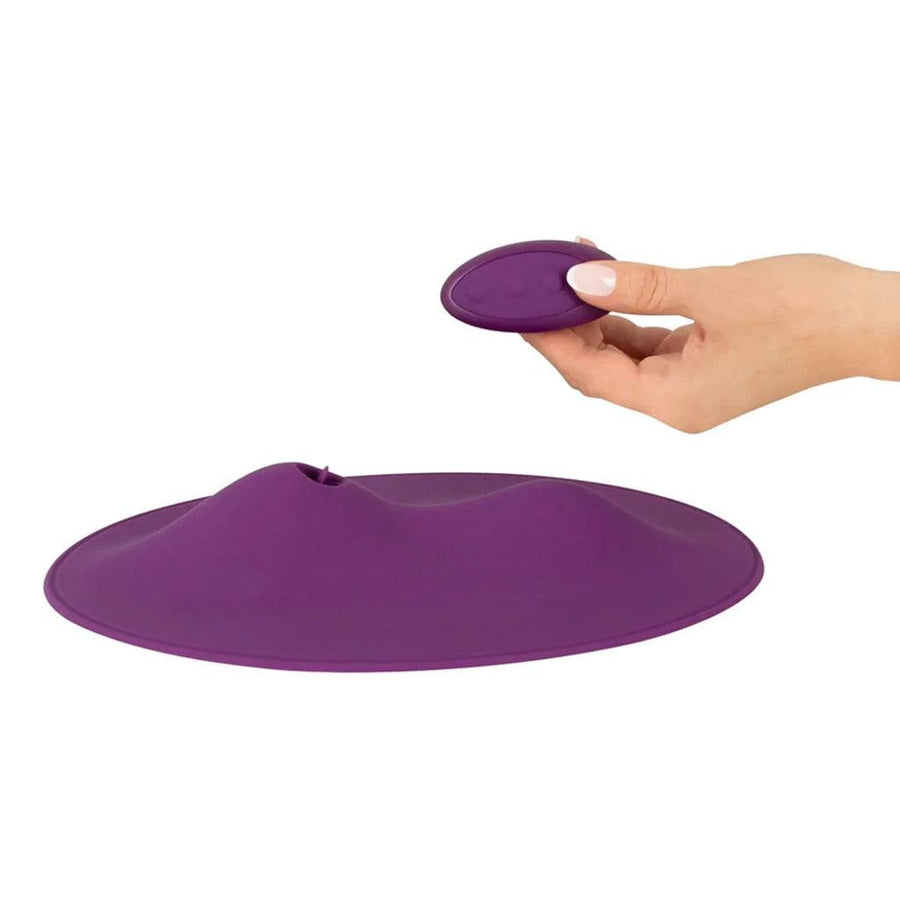 The Vibepad 2 Remote Controlled Grinding Vibrator Pad is shown against a blank background. A hand is shown holding the remote, which has three buttons and is the same color as the vibrator.