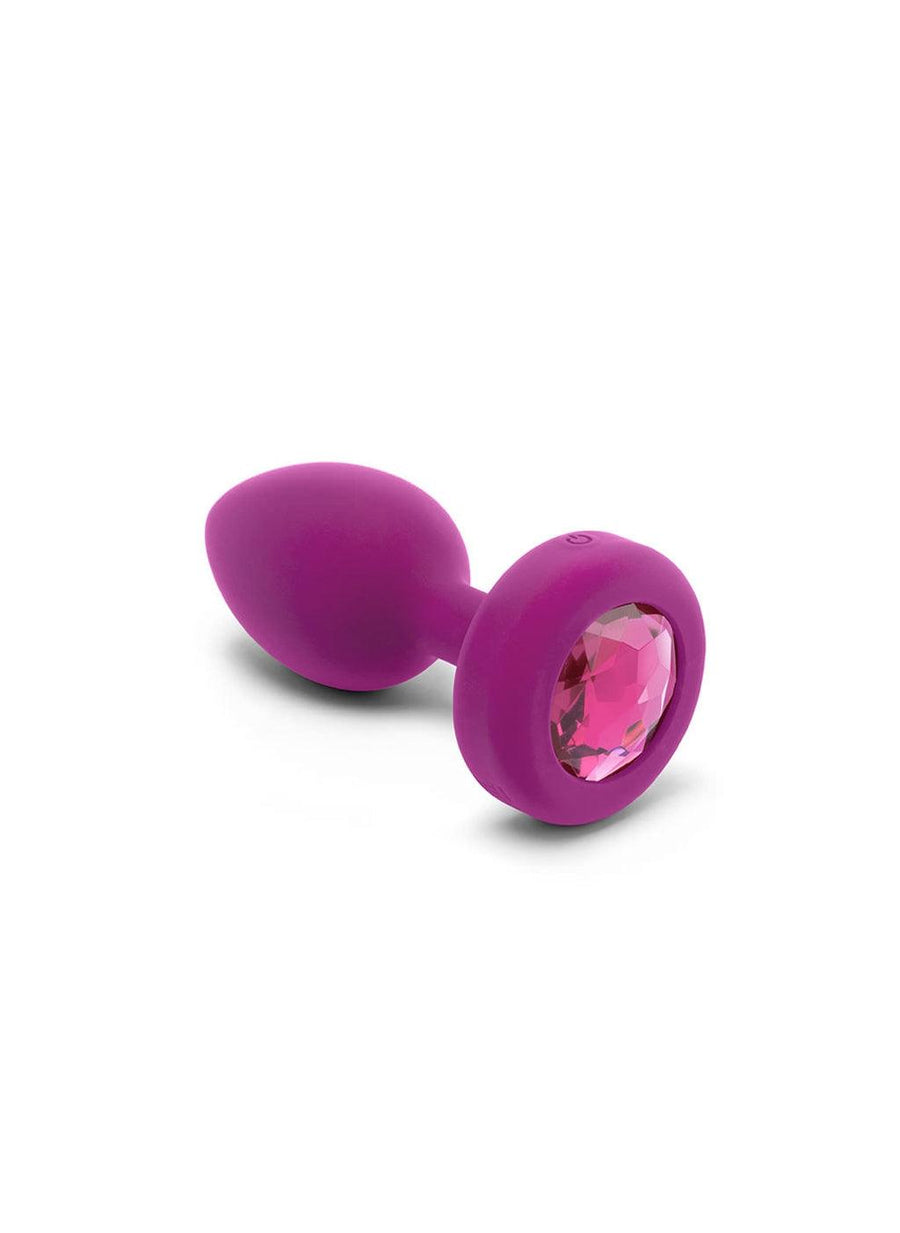The b-Vibe Vibrating Jewel Butt Plug in Fuchsia is shown sideways against a blank background. Its power button is visible on the base, and the back of the base has a large, fuchsia crystal embedded in it.
