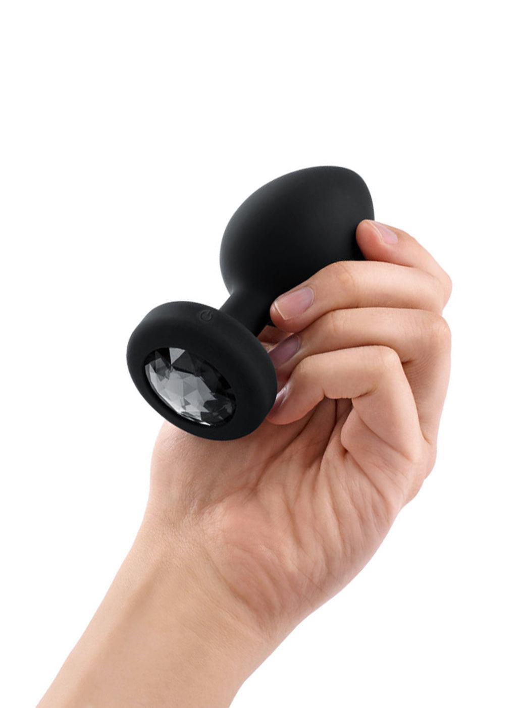 A hand is shown holding the b-Vibe Vibrating Jewel Butt Plug in Black against a blank background on its side, displaying the black crystal on the base.