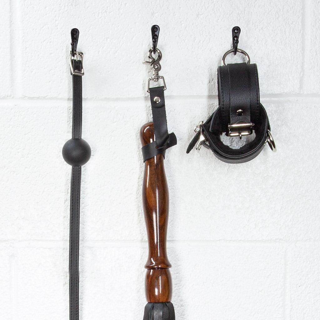 A close-up image of the Sissoo Rosewood Long Handle Leather Flogger with leather hanging strap on a white background. The flogger is shown hanging on a black hook next to leather wrist cuffs and a black silicone ball gag made by The Stockroom.