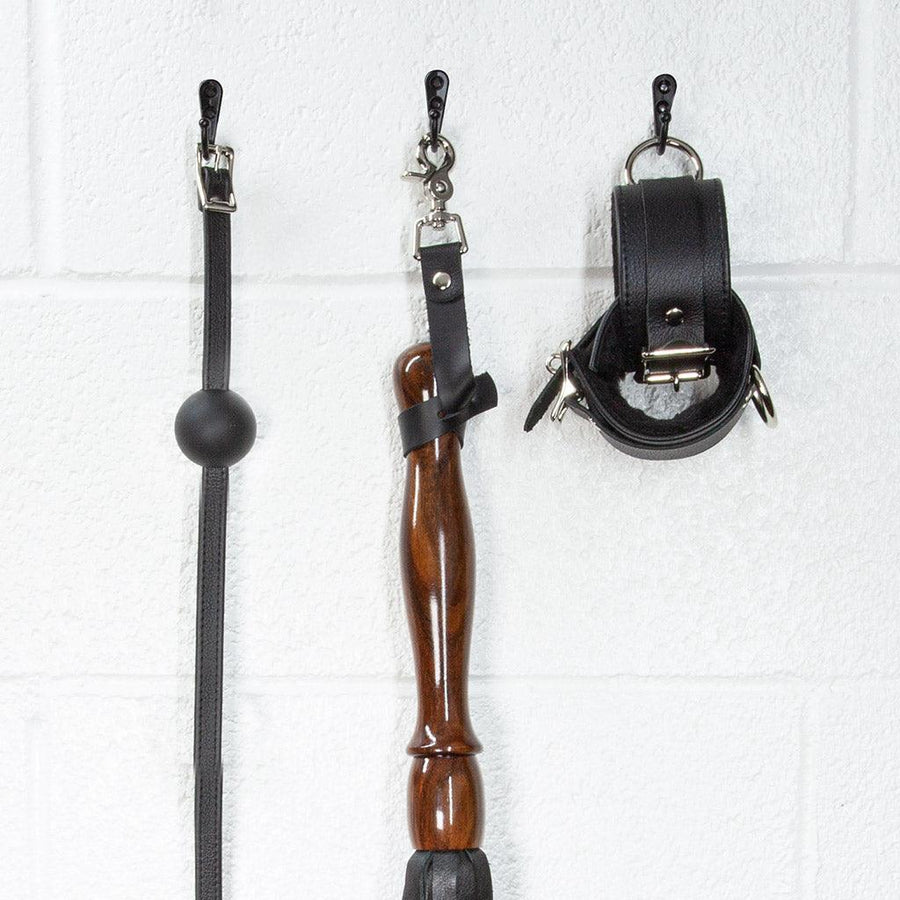 A close-up image of the Sissoo Rosewood Long Handle Leather Flogger with leather hanging strap on a white background. The flogger is shown hanging on a black hook next to leather wrist cuffs and a black silicone ball gag made by The Stockroom. 