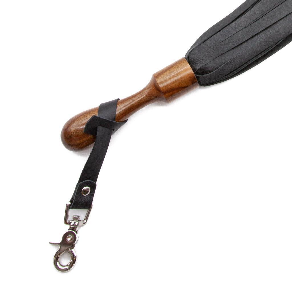 A close up image of the Sissoo Rosewood Short Round Handle Leather Flogger on a white background. Shown is the included leather hanger strap with crab-claw hook attached to the handle of the flogger.