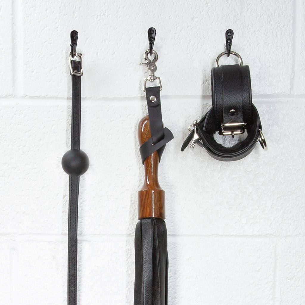 A close-up image of the Sissoo Rosewood Short Handle Leather Flogger with leather hanging strap on a white background. The flogger is shown hanging on a black hook next to leather wrist cuffs and a black silicone ball gag made by The Stockroom.