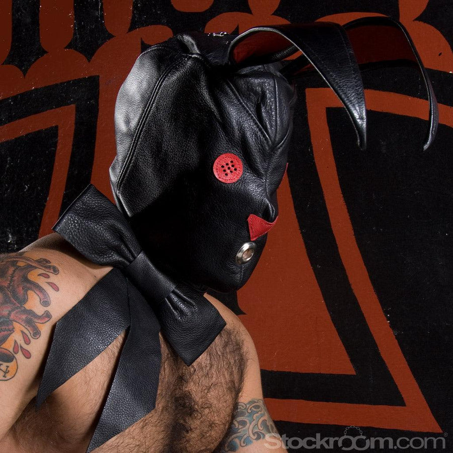 A man with tattoos and dark chest hair stands in front of a red wall with a black design on it. He is wearing the black Leather Bunny Hood.