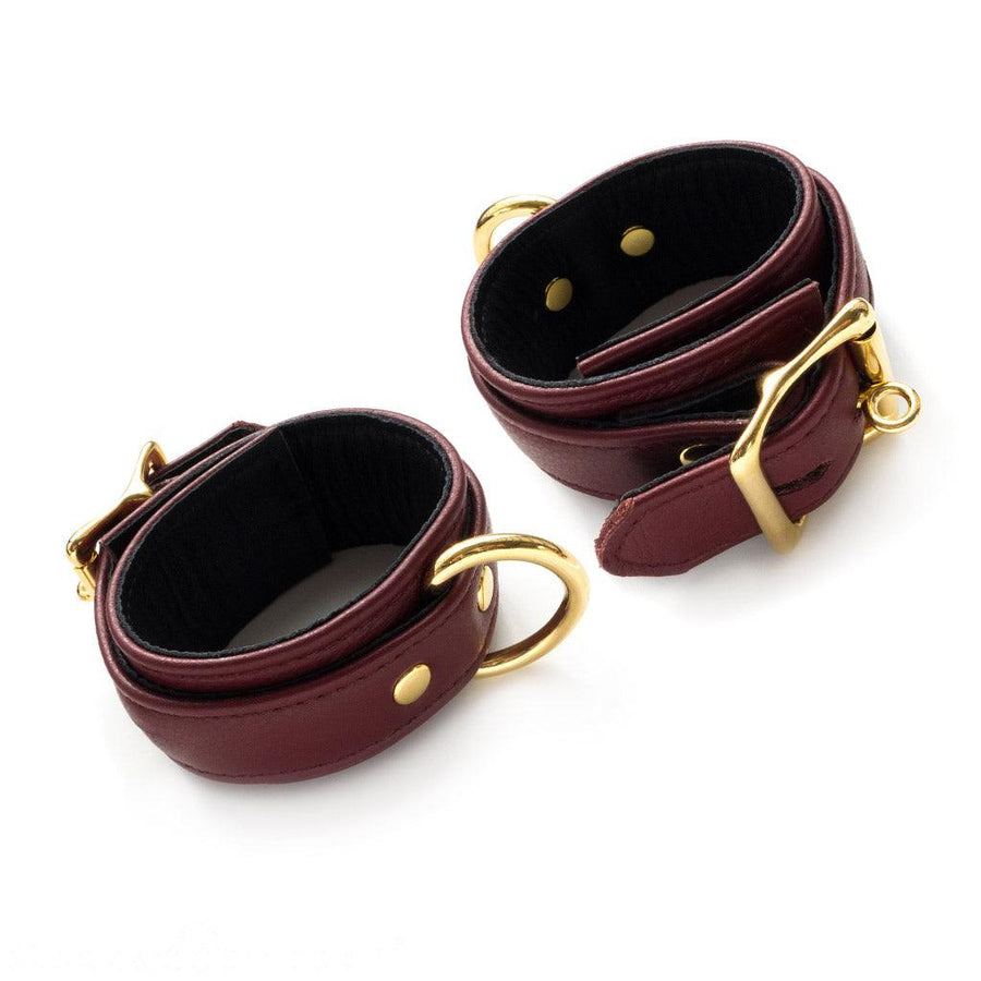 The JT Signature Collection Wrist Restraints are shown against a blank background. The cuffs are made of Bordeaux leather with a black interior lining. The cuffs have a D-ring and a buckle closure. The hardware is gold.