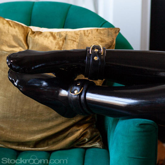 Somebody’s lower legs, clad in black latex stockings, are shown resting on the arm of a teal velvet chair, displaying the black Garment Leather Ankle Cuffs With Brass Gold Hardware that they are wearing.