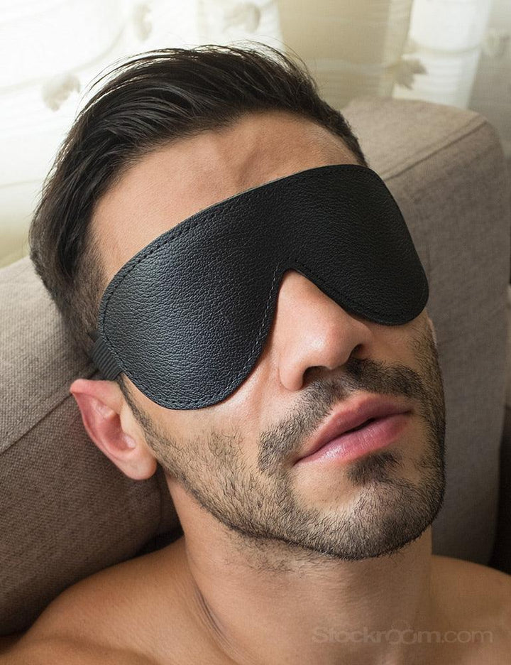 A brunette man with facial hair leans his head back against a grey couch. A close-up of his face is shown, and he wears the Padded Leather Blindfold with an Elastic Band. The blindfold fits his face and is made of black leather.