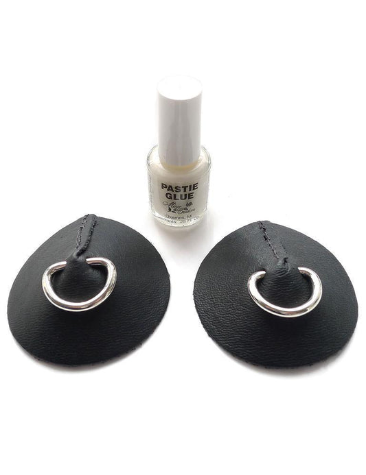 The Black Leather Pasties with D-rings are displayed against a blank background alongside a bottle of pastie glue. The pasties are black leather circles and have metal D-rings in the center, resembling nipple piercings.