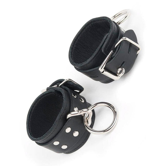 A pair of black Premium Leather Cuffs With A Locking Buckle is displayed against a blank background. The cuffs have a lockable metal buckle and a metal triangle, which has an O-ring dangling from it.