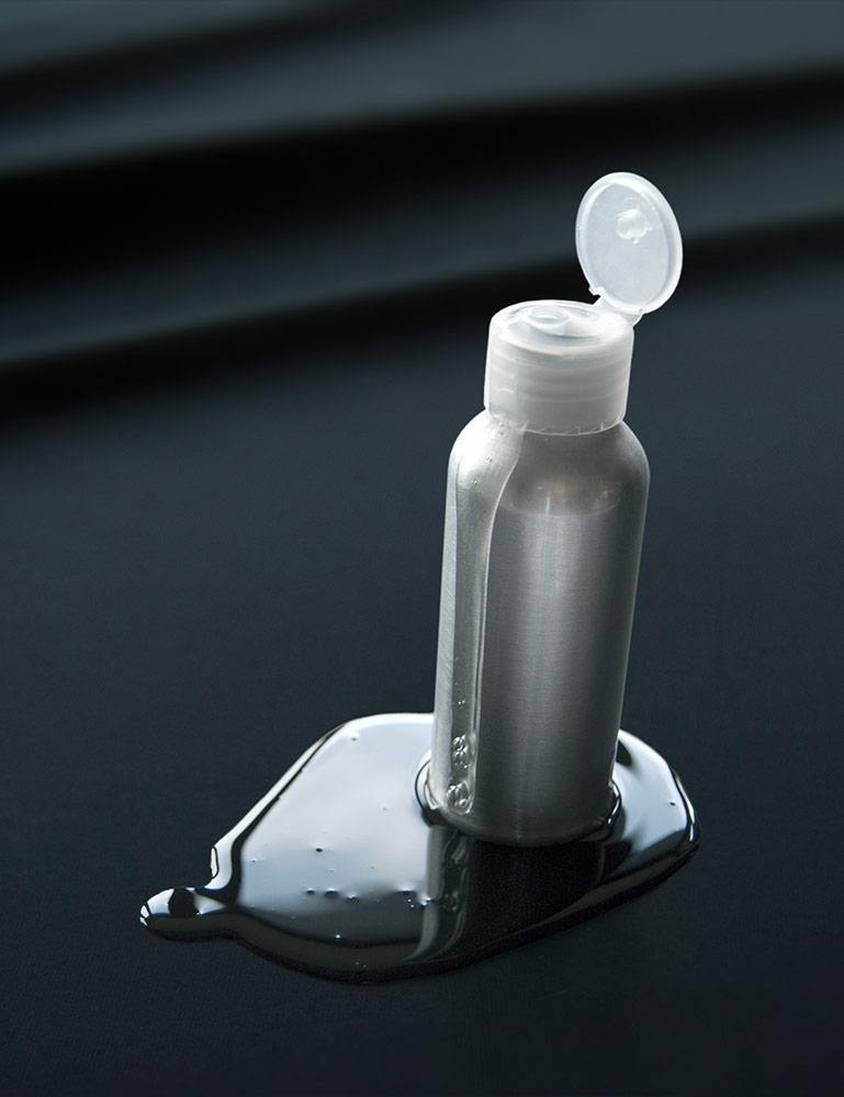 A silver bottle of lube with a flip-cap is shown standing on the Fluid Proof Protective Throw By Sheets Of San Francisco. There is a pool of lube under the bottle, but the fluid rests on top of the sheet instead of soaking into it.