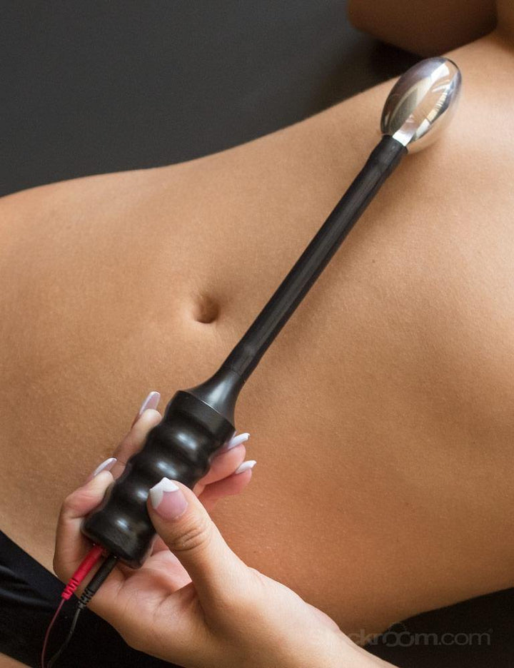 A close-up of a woman’s bare stomach is shown against a grey background. She has a French manicure and holds the Electrastim Depth Charge Electro Dildo on her stomach.