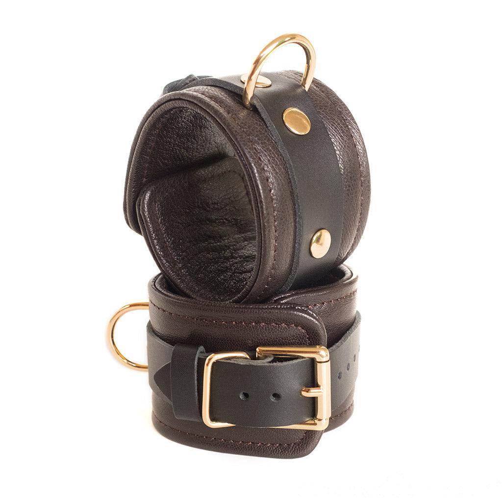 The Brown Leather Wrist Restraints With Gold Accent Hardware are shown stacked on top of each other against a blank background. They are fastened with adjustable buckles.
