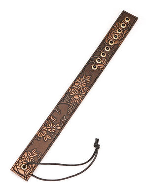 The Vegan Floral Ruler With Gems is shown against a blank background. It is a piece of brown faux leather shaped like a ruler with a floral design in light brown. There is a row of black gems at the top of the ruler and a wrist loop at the bottom.
