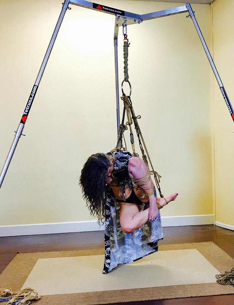 A woman is shown suspended from the Tetruss Maxximus Suspension Bondage Frame. She is suspended by her leg, which is tied in a bent position. Her brown hair hands down as she lowers her head.