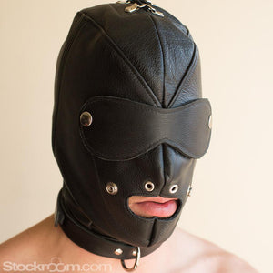 A person's head in the Premium Leather Hood with a Gag and Blindfold is shown against a beige background. The hood is made of black leather with silver hardware. The gag has been removed, exposing their mouth. The blindfold is hourglass-shaped.