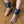 Load image into Gallery viewer, A close-up of a man&#39;s lower legs is shown with his feet resting on a wooden floor. He wears the Premium Lockable Dungeon BDSM Black Leather Ankle Cuffs. The cuffs are wide and have metal hardware. They are connected with a metal snap hook.
