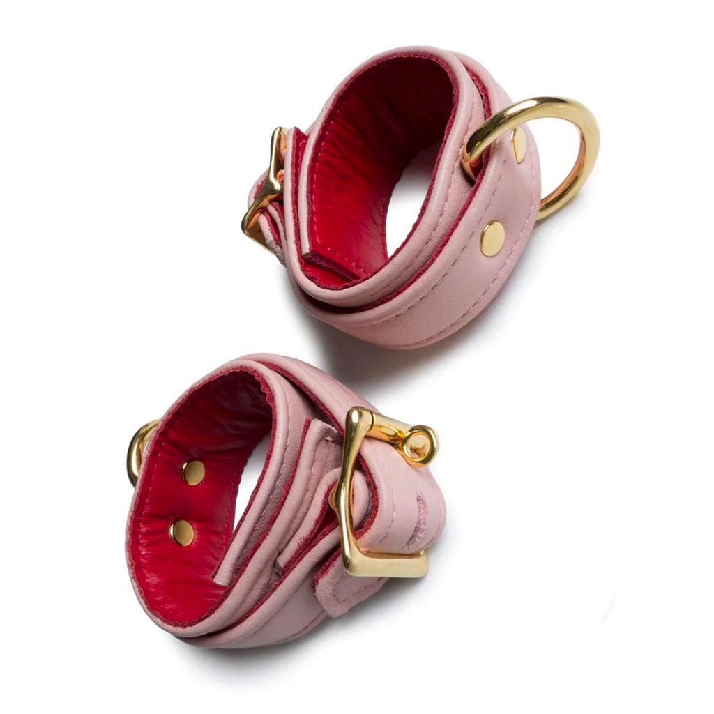 A pair of Premium Garment Leather Wrist Cuffs With 18 Karat Gold Plated Hardware is shown against a blank background. The cuffs are light pink with red interior lining. The buckles, D-rings, and rivets are all gold.