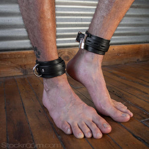 A close-up of a man’s bare feet standing on a wooden floor is shown. There is a metal wall in the background. He has a pawprint tattoo on his lower leg and is wearing the black Premium Garment Leather Ankle Cuffs. They are locked with small silver padlocks.