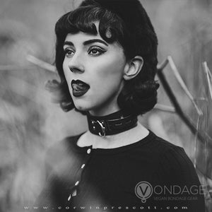 A black and white photo shows a woman with 50s style dark hair and clothes standing in a field. She wears a dark buttoned-up cardigan and beret, as well as the Vondage Locking/Buckling Collar. She is licking her lips.