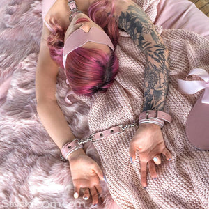 A woman with pink hair wearing a pink latex bra lays on a bed with pink blankets. Her arms are above her head, and the Stupid Cute Restraint Clip attaches her matching handcuffs. She also wears a matching blindfold and a bust harness.