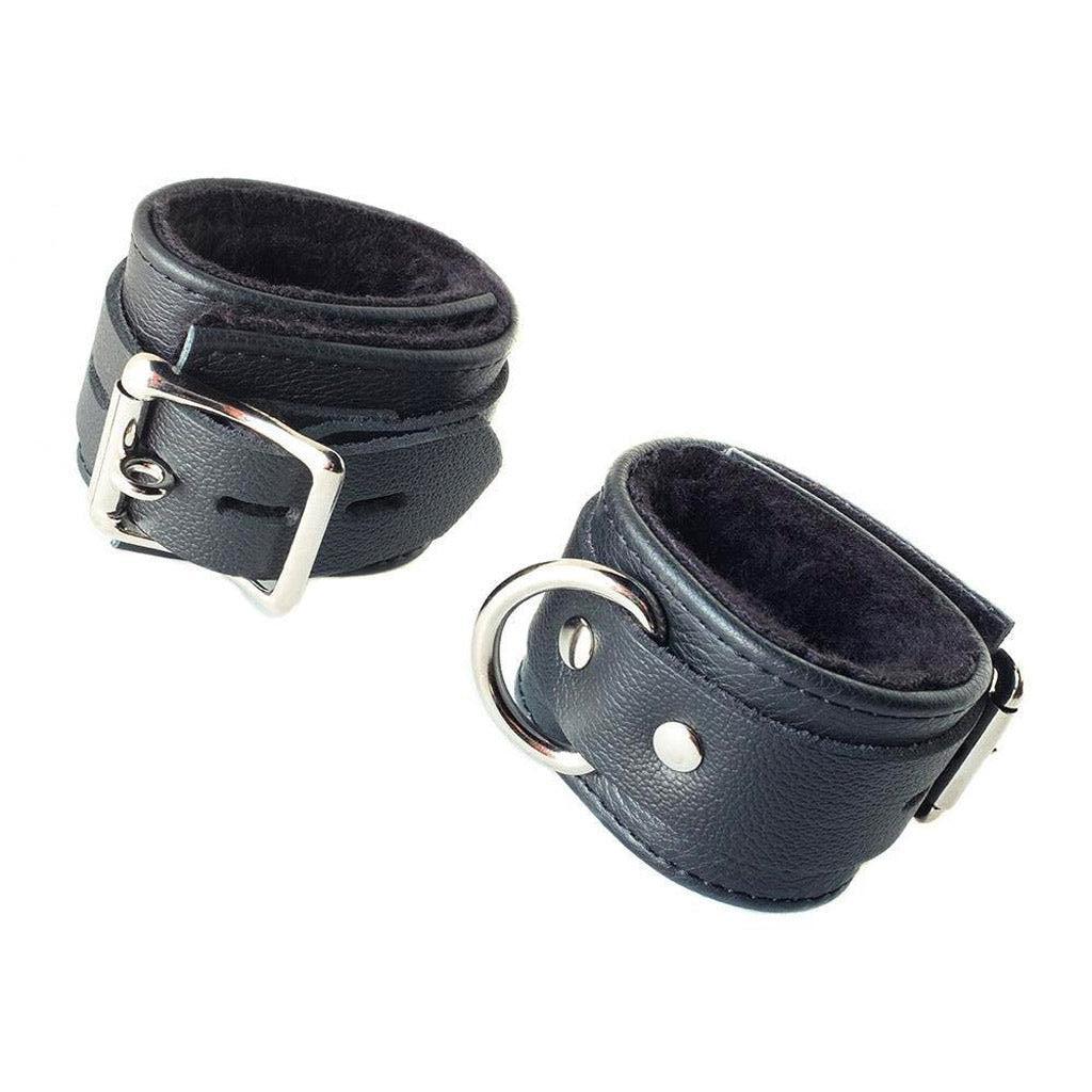The Fleece Lined Garment Leather bondage Wrist Cuffs are shown cuffed against a blank background. The cuffs are made of black leather and have silver hardware. The cuffs each have a D-ring and a buckle. The inside of the cuffs is lined with black fleece.
