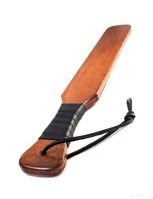 The Leather Wrapped Narrow Wood Spanking Paddle is shown against a blank background. The paddle is made of light-colored wood in the shape of a rectangle with a thinner handle. The handle is wrapped in black leather and has a wrist loop.