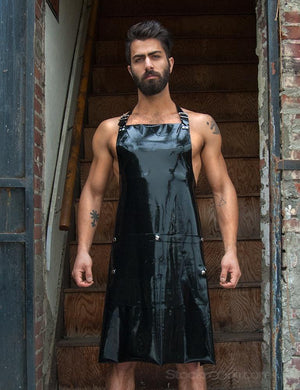 A muscular brunette man with facial hair stands in front of a wooden staircase. He wears the black Rubber Apron by Syren Latex, which has pockets and an adjustable neck strap. The hem of the apron ends at his knees.