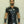 Load image into Gallery viewer, A brunette man with facial hair poses in front of a white wall. He wears the Uniform Shirt from Syren Latex in black, matching pants, and arm gauntlets. The shirt has metal snaps down the front, pockets on the chest, and a collar.
