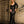Load image into Gallery viewer, A blonde woman poses in front of a brick wall next to a large metal cage. She wears the Madonna Latex Gown by Syren Latex in black. The gown has a low neckline and long sleeves. It is fitted until the knees, where it flares out.
