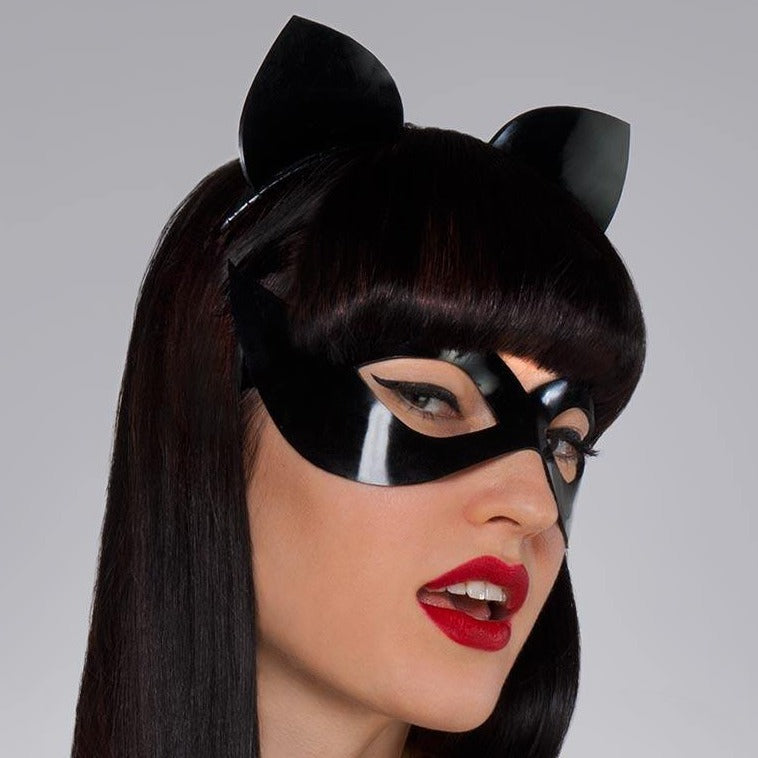 A headshot of a woman with dark hair and red lipstick shot against a grey backdrop. She wears the Kitten Headband in black by Syren Latex. She also wears a black latex mistress mask.