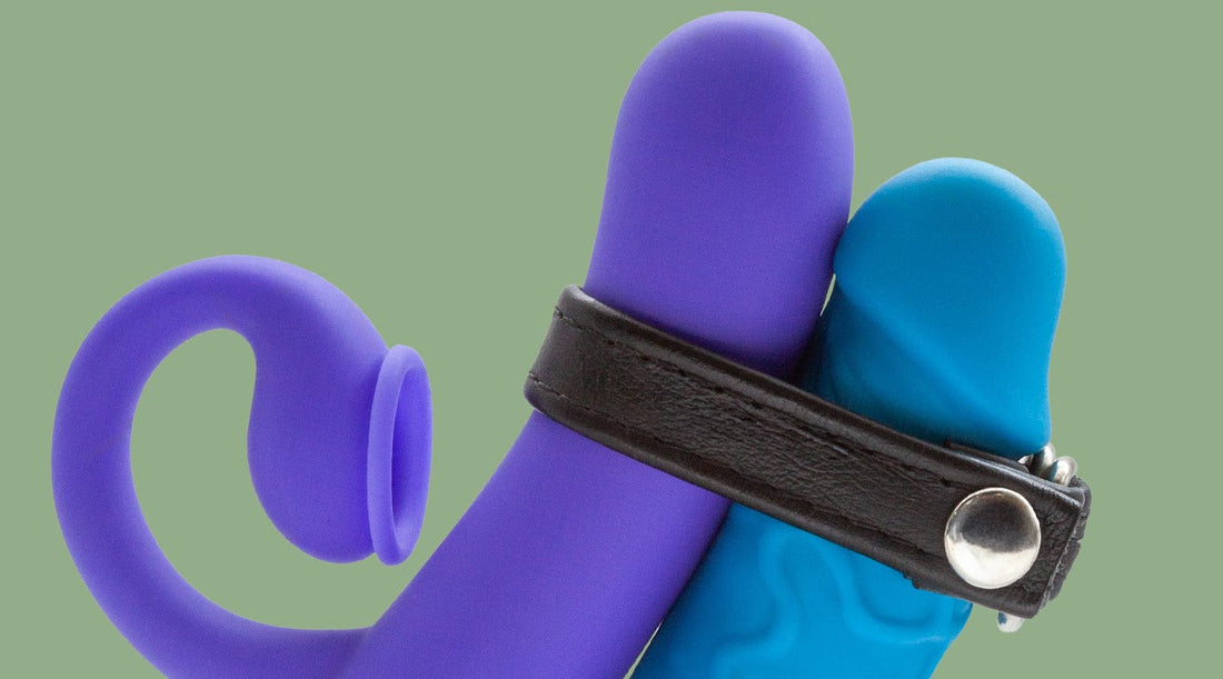 A purple vibrator held against the shaft of a blue dildo with a black leather C-ring.