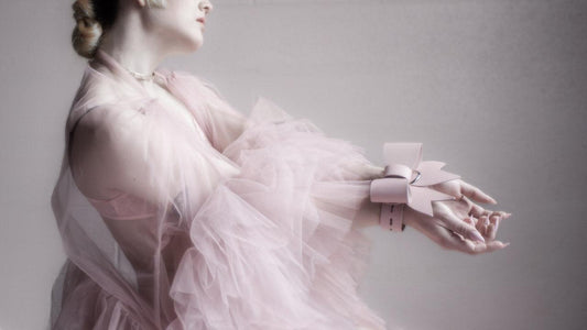A woman in a gauzy pink dress stands against a grey background, holding out her hands. Her wrists are fastened together with the Stupid Cute Bow Restraint from Stockroom. 