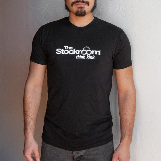 A man is shown wearing a Men's Stockroom Think Kink T-Shirt. The T-shirt is black and has the Stockroom logo printed in white on it, with the slogan “Think Kink” underneath.