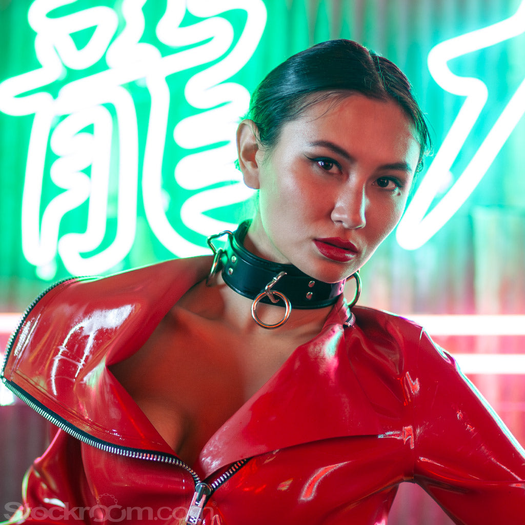 A woman with dark hair sits in front of a glowing green and red neon sign. She is wearing a red latex moto jacket and black latex panties. She is wearing the black leather 3-Ring Slave Collar With A Locking Buckle.