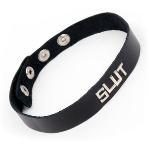 The Slut Black Leather Word Collar is displayed against a blank background. It is a thin strip of black leather with the word slut on the front in silver letters. The collar has snap closures in the back.