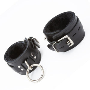 A pair of black Premium Fleece-Lined Leather Cuffs With A Locking Buckle is displayed against a blank background. The cuffs have a lockable metal buckle and a metal triangle, which has an O-ring dangling from it. The cuffs are lined with black fleece.