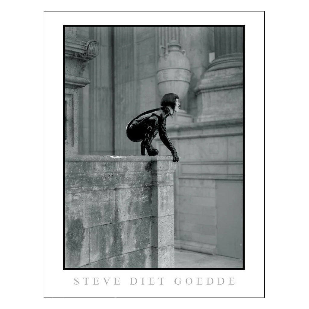 A poster of a Steve Diet Geodde black and white photograph Of Mistress Midori dressed in black latex perched on the corner of a concrete Roman style building is shown on a white background. 