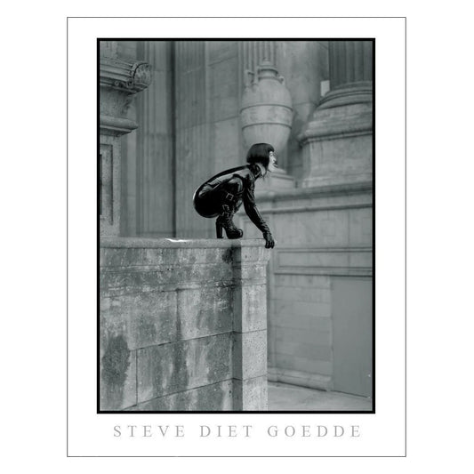 A poster of a Steve Diet Geodde black and white photograph Of Mistress Midori dressed in black latex perched on the corner of a concrete Roman style building is shown on a white background. 