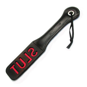 The Slut Impression Paddle is displayed against a blank background. The paddle is rectangular with a wrist loop and made of black leather with the word slut cut out, revealing red leather underneath.