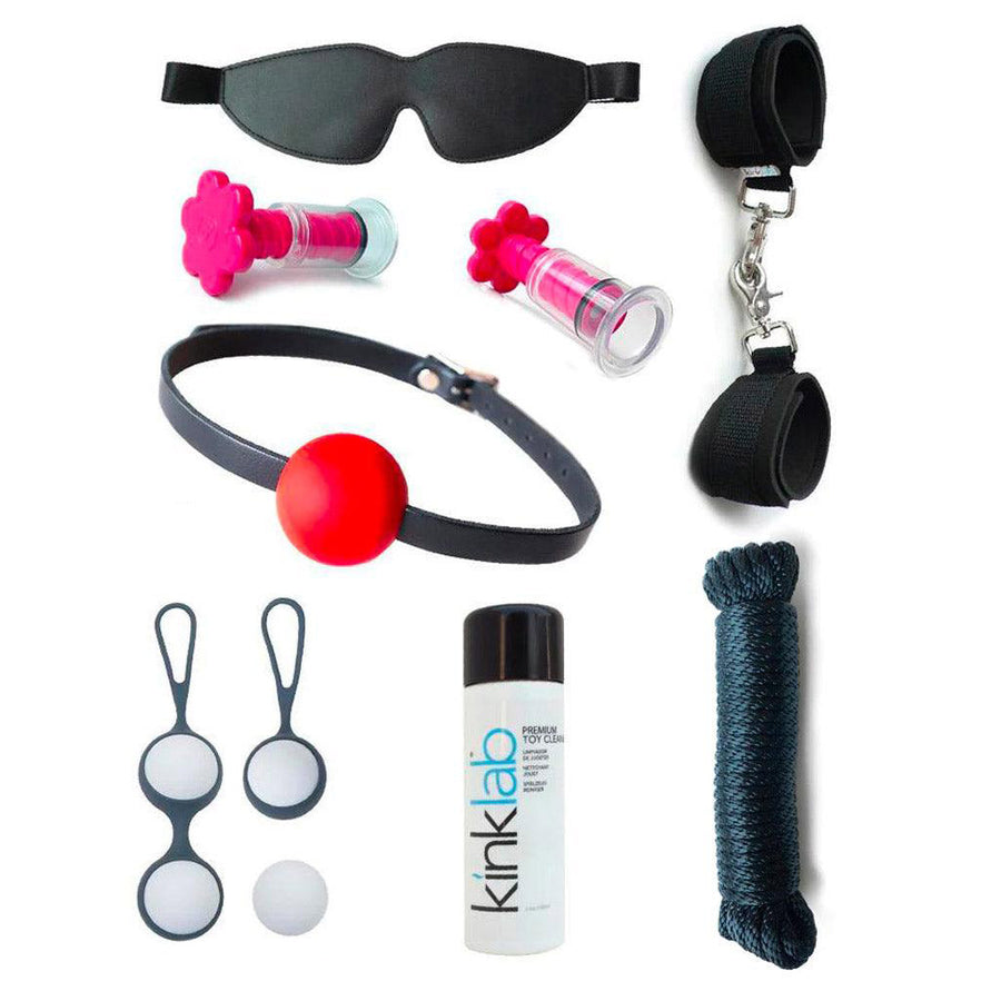 The contents of the Deluxe Stay At Home Adventure Kit are shown against a blank background. Shown is a black blindfold and wrist cuffs, black rope, pink suction T-cups, toy cleaner, a ball gag, and kegel balls.