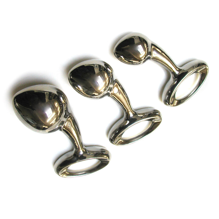 A small, medium, and large Njoy Pure Plug are laid next to each other against a blank background. They are made of polished stainless steel and have a tapered, conical shape. The neck of the plug is slightly curved, and the base is a large ring.