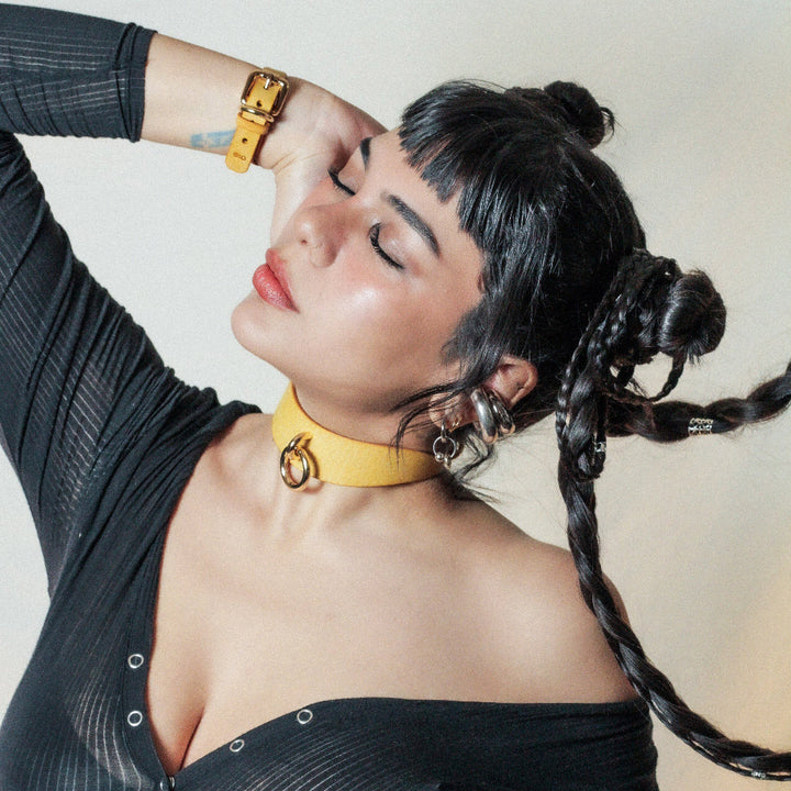 A young woman with short black bangs and long braids tilting her head to the right of the viewer. She's wearing the Orbit Day Collar by Neon Coyotes on her throat, in "Golden Hour," a bright yellow color of leather, with gold-colored hardware. 