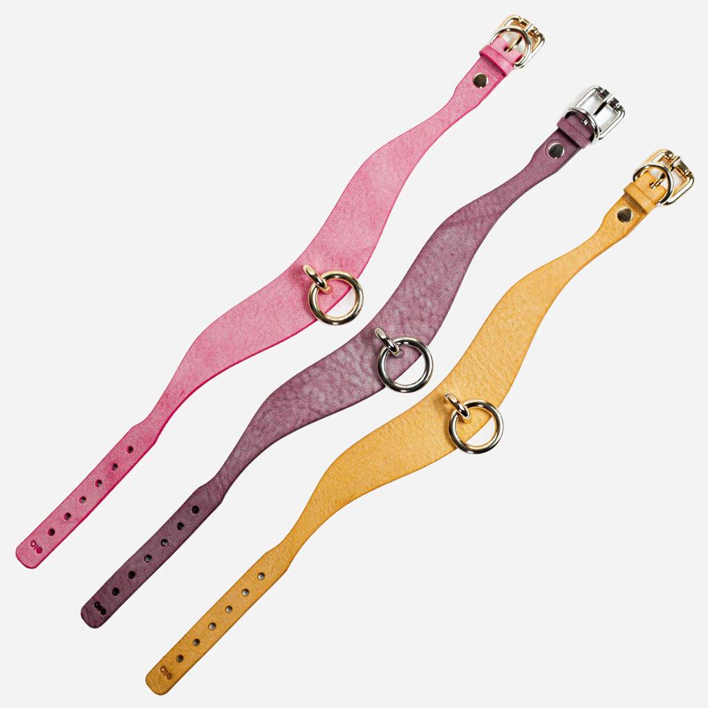 Three models of the Orbit Day Collar from Neon Coyotes laid out flat and diagonal on a white background. From top to bottom, the colors are: Blush, a rosy pink color with gold-colored nickel hardware; Haze, a smoky lavender color with silver-colored nickel hardware; and Golden Hour, a bright yellow leather with nickel hardware in a gold tone. 