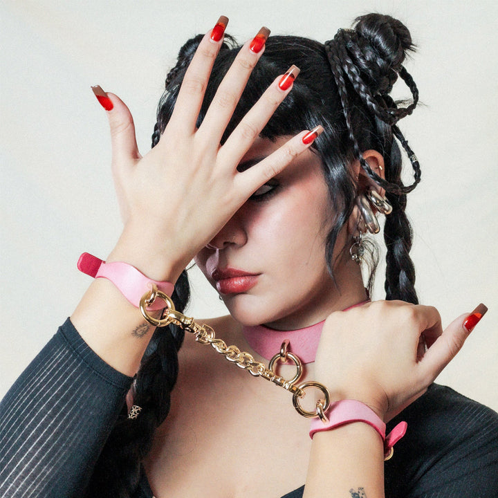 A young woman with black hair spreading her hand in her face. Her wrists are bound with pink leather BDSM cuffs and a gold-colored chain linking them. 