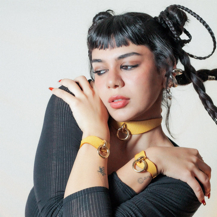 A young woman with short black bangs and long braids, crossing her hands over her chest. Her wrists have bright yellow leather BDSM cuffs on them.